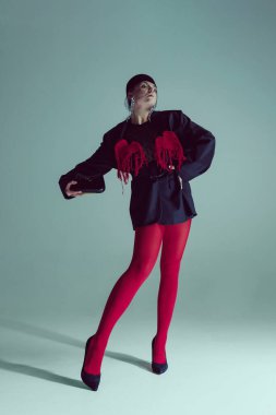 Siyah ceketli, kırmızı taytlı, küçük el çantası tutan, stüdyo arka planına karşı açılı pozlar veren kendine güvenen genç bir kadın. Yenilikçi moda anlayışı, zarafet, cesur sürrealizm, yüksek moda.