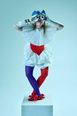 Renkli kıyafetleri olan, kırmızı korseli, mavi şapkalı, beyaz arka planda dürbün olarak zilleri tutan biri. Gerçeküstü moda kavramı, güzellik, çeşitlilik, kendini ifade etme, sanat. Ad