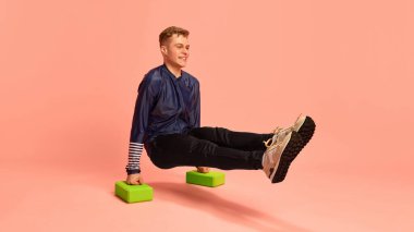 Genç adam mavi atletik bluz ve siyah pantolon giyen köpük bloklarda yüksek L-sit performansı sergiliyor. Pembe arka plana karşı dayanıklılık ve çekirdek gücü gösteriyor. Fitness kavramı, denge, çekirdek egzersizi.