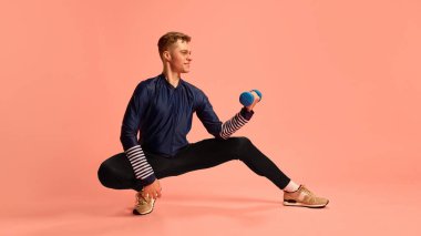 Geniş hamlede spor erkek, pazı kıvrımlı dambıl tutuyor, mavi spor kıyafeti giyiyor, pembe arka planda çalışırken gülümsüyor. Güç kavramı, eğitim, fiziksel aktivite, beden eğitimi.