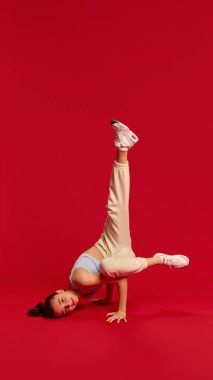 Hafif giysili genç bir kız tek koluyla dengede duruyor, bacağı yukarıda, kırmızı arka plandaki egzersiz sırasında güç ve kontrol gösteriyor. Spor anlayışı, fiziksel uygunluk, beden dengesi.