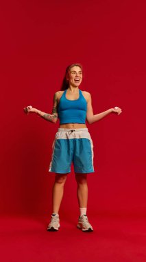 Mavi üniformalı, neşeli yüzlü, kırmızı stüdyo arka planında biceps kıvrımları yapan zayıf genç bir kadının tam vücut fotoğrafı. Spor, spor, sağlık, beden eğitimi kavramı. Ad