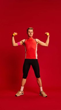 Kırmızı kolsuz gömlekli, turuncu halterli, güçlü poz veren, spor yaparken kaslarını ve gücünü gösteren kendine güvenen bir adam. Vücut gücü, ağırlık eğitimi, sağlık kavramı.