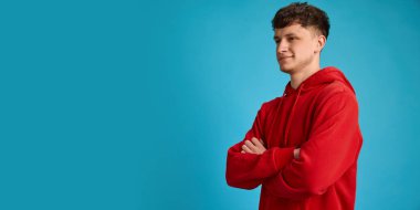 Kırmızı kapüşonlu genç adam kollarını kavuşturup sırıtarak mavi stüdyo geçmişine bakıyor. Güven, yaşam tarzı, kişilik, nüfuz, karizma, gündelik moda kavramı