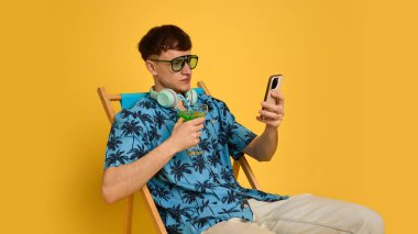 Ciddi bir adamın elinde içki ve telefonla oturmuş, odaklanmış bir ifadeyle göz gezdirdiği orta boy bir fotoğraf. Uzak çalışma, tatil, dijital yaşam tarzı, iletişim, internet, gençlik kavramı.