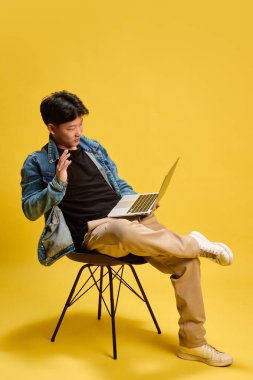Dizüstü bilgisayarlı genç adam gülümsüyor ve online haberleşme jestinde elini kaldırıyor, sandalyede oturuyor. Sanal toplantı kavramı, iş ağı, uzak çalışma, markalaşma, güven, teknoloji dünyası.