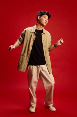 Kendine güvenen, bej kıyafetli, kırmızı arka planda havalı bir şekilde dans eden genç adamın uzun bir fotoğrafı. Tarz, ritim, gençlik kültürü, özgürlük, kendini ifade etme, internet, nesil, neşe kavramı.