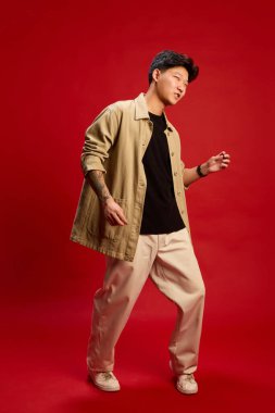 Gündelik bej kıyafetli gülümseyen genç adam kırmızı zemine karşı dans hareketlerinde güvenle hareket ediyor. Kutlama, ifade, yaşam tarzı, yaratıcılık, moda ileri kişiliği kavramı.