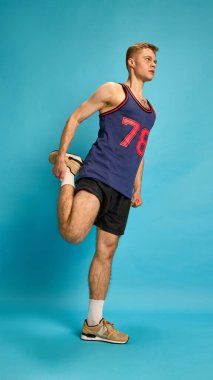 Atletik genç bir adam tek ayak üzerinde durarak bacaklarını esnetiyor, esneklik ve ısınma süreci gösteriyor, stüdyo arka planına karşı. Antrenman öncesi rutin, fiziksel bakım ve günlük hazırlık kavramı.