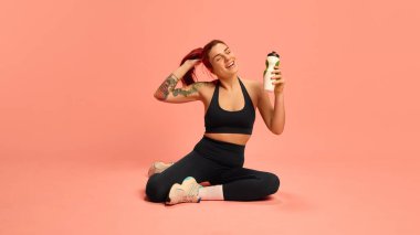 Siyah spor kıyafetli rahat bir kadın bacak bacak üstüne atmış, elinde su şişesi, şakacı bir şekilde saç kesme egzersizi yapıyor. Spor, su, iyileşme, sağlık, fiziksel sağlık kavramı.