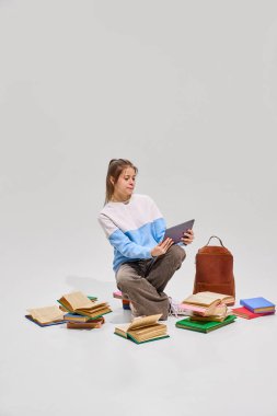 Kitaplara odaklanmış, etrafı kitaplarla çevrili, elinde tablet olan ve beyaz stüdyo geçmişine karşı öğrenen genç bir kız. Eğitim, lise, diploma, öğrenim ve öğrenim kavramı. Ad