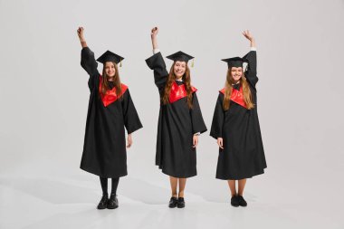 Genç, üniversite mezunu kızların toplu portresi, siyah elbiseler ve şapkalar giyip, gri arka plana karşı neşe ve mutlulukla el kaldırıyorlar. Eğitim anlayışı, lise, diploma, kutlama.