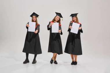 Siyahi elbiseler ve şapkalar giymiş üniversite mezunu genç kızların toplu portresi, sertifikalarını gösteriyor ve gri arka planda gülümsüyor. Eğitim anlayışı, lise, diploma, eğitim. Ad