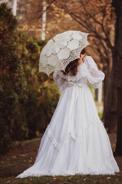 Altın sonbahar parkında dantel şemsiyenin altında prenses gibi görünen, dalgalı beyaz elbiseli Viktorya dönemi kadını. Eski moda, nostalji, rüya manzarası, güzellik. Film ve kitap kapakları için uygun.