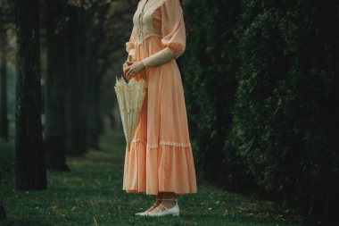Şeftali rengi elbisesi, kapalı şemsiyeyle hareketsiz beklerken ıslak çimenlere değiyor. Varlığı, ormandan yükselen bir anı gibi ruhani. Sessizlik kavramı, melankolik. Kitap veya film kapağı için uygun.