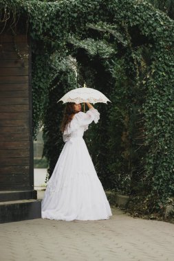 Eski moda beyaz elbiseli ve şemsiyeli zarif bir kadın yeşil kemerin altında yukarı bakıyor. Umut kavramı, hayal, sonsuz zarafet. Romantik edebiyat ya da klasik drama için uygun.