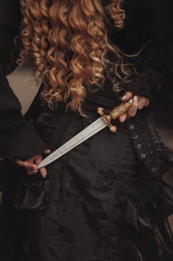 Siyah korse ve dalgalı buklelere karşı altın hançer tutan genç bir kadının detaylı görüntüsü. Klasik, intikam, miras, unutulmuş kraliçeler kavramı. Gotik kurgu ya da tarihi intikam öyküsü için uygun..