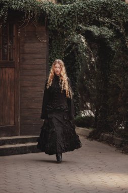 Siyah dantel cüppeli altın bukleli genç kadın ahşap evin yanındaki sarmaşık kemerinin altında yavaşça yürüyor. Gizem, gotik güzellik ve sessiz varlık kavramı. Karanlık peri masalı ya da roman kapağı için uygun.