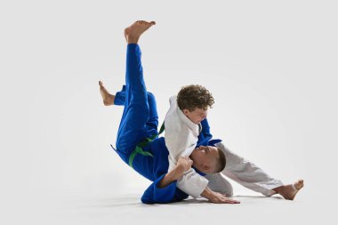Beyazlı genç judoka, yoğun zemin kontrolü sırasında mavili rakibini tutuyor. Kontrolü ve beyaz arka plana karşı gücünü gösteriyor. Zafer kavramı, dövüş sanatları, dövüş sporları, yarışma.