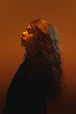 Yumuşak kıvırcık saçlı, ifadesiyle turuncu arka planda poz veren bir kadının yan profili. Güzellik kavramı, moda, sessizlik duygusu, kimlik, ruh hali, kendi yansıması. Ad
