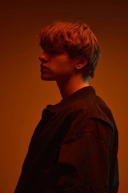 Profilde genç bir adam, siyah ceket giyiyor, turuncu ışık altında sıcak turuncu stüdyo arka planına ciddi ciddi bakıyor. Güç kavramı, durgunluk, iç yansıma, cesur erkeklik, duygu.