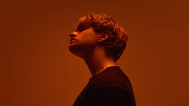 Kafası eğik, yumuşak turuncu sisli parlak eğimli arka planda okunamayan genç adamın yan görüntüsü. Güzellik kavramı, sakinlik, duygusal kontrol, sessiz drama, kendini tanımlama..