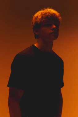 Gölgelerin içindeki genç adam, yüzü kısmen gizlenmiş, sıcak dramatik neon ışıklarıyla sıcak turuncu arka planda. Güzellik kavramı, moda, erkeksi, duygusal ihtiyat, sessiz gerilim.