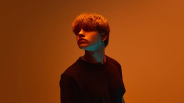 Genç adam, sakin ve odaklanmış bir ifadeyle, turuncu rengin altında güçlü bir kontrastlı ışık altında başını yana çeviriyor. Güzellik ve gençlik kavramı, duygu, moda, modern. Ad