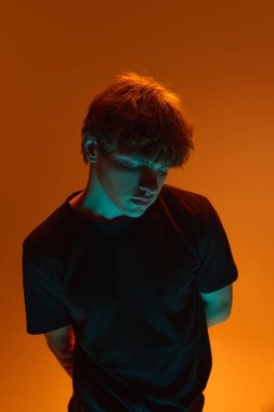 Yaratıcı neon portresi, gözleri aşağıda, kolları arkada turuncu neon ışıkta duran, parlak saçlı genç bir adam. Güzellik ve moda kavramı, kendini ifade etme, insani duygular.