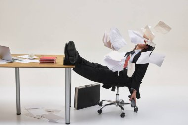 Masanın üzerinde bacakları olan sandalyede oturan bir adam kaotik ofis karmaşasında uçuşan kağıtlarla çevrili, işten bunalmış. Dinamik fotoğraf. Ofis hayatı kavramı, baskı, çoklu görev, iş kaosu. Ad