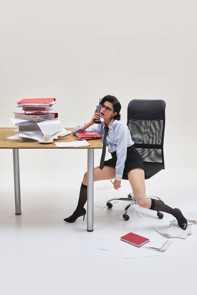 Молодая женщина разговаривает по телефону, сидя сутулившись в открытой позе, окруженная офисным беспорядком и незаконченными заданиями. Концепция офисного образа жизни, разъединения, выгорания, бизнеса.