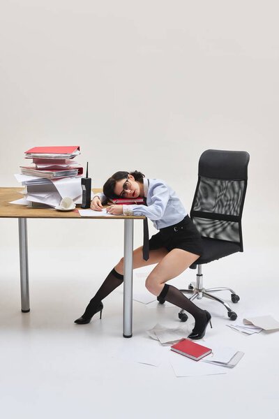 Молодая женщина лежит головой на документах, опираясь на стол с сонным выражением лица в грязной офисной обстановке на белом фоне студии. Концепция офисного образа жизни, усталости, выгорания, бизнес-давления.