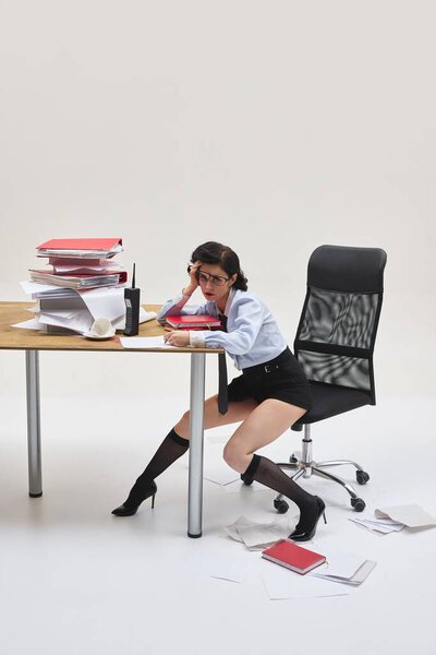 Молодая женщина, присевшая рядом с столом, сложенная из папок, держась за голову, в то время как переполнена стрессом. Концепция офисного образа жизни, тревоги, рабочего давления, документации.