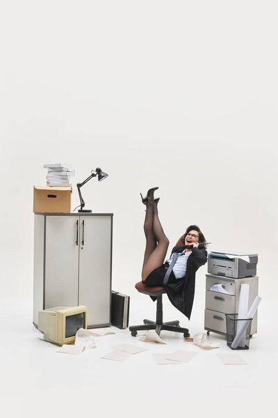 Молодая женщина смеется по телефону с ногами в воздухе, сидя в грязном углу офиса, окруженный бумажной работой и устройствами. Концепция офисного образа жизни, человеческих эмоций, социальных проблем, бизнеса.