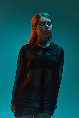 Saf bluz giymiş genç bir kadın, temiz stüdyo arkaplanına karşı neon deniz mavisi ışığında sakin bir şekilde poz veriyor. Güzellik ve moda kavramı, kendini ifade etme, insani duygular.