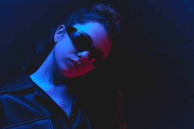 Güneş gözlüklü genç bir kadının yakın plan portresi mavi ve kırmızı ışık altında koyu arkaplanda siyah deri kıyafetler giyiyor. Güzellik ve moda kavramı, kendini ifade etme, insani duygular.