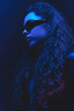 Fütürist güneş gözlüklü ve parlak neon ışıklı deri ceketli genç bir kadın, karanlık stüdyo arka planında. Güzellik ve moda kavramı, kendini ifade etme, insani duygular.