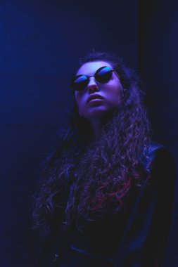 Şık, yuvarlak güneş gözlüklü, uzun kıvırcık saçlı ve koyu arka planda neon yansımalı kadın portresi. Güzellik ve moda kavramı, kendini ifade etme, insani duygular.
