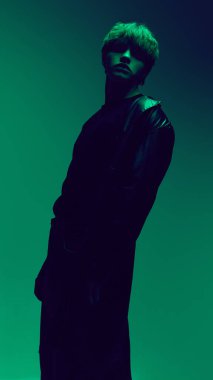 Moda modeli yeşil ışıkta etkileyici bir şekilde poz veriyor. Tarz sahibi siyah bir ceket giyiyor ve arka planda koyu bir ifade sergiliyor. Güzellik ve moda kavramı, kendini ifade etme, insani duygular.