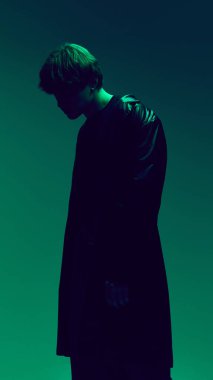 Koyu yeşil stüdyo ışığı altında, arka planda esrarengiz bir ruh hali olan ceketli genç adam silueti. Güzellik ve moda kavramı, kendini ifade etme, insani duygular.