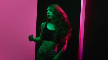 Modaya uygun deri ceketli genç bir kadın, çarpıcı pembe neon ışığı ve koyu arkaplana karşı yeşil alçı altında yoğun bir şekilde bakıyor. Etkileyici bir stüdyo resmi. Güzellik ve moda kavramı, kendini ifade etme