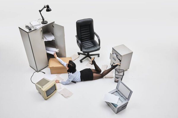Фото с воздуха. Женщина, лежащая и залезающая в кабинет в окружении бумаги и беспорядка в нереальной белой офисной сцене. Концепция усталости, нереалистичные требования, женская роль в корпоративной структуре.