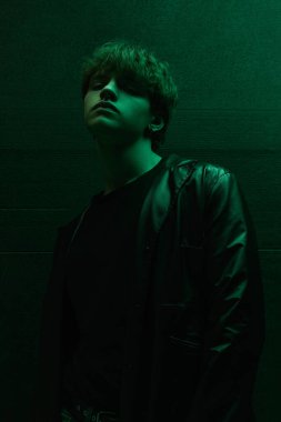 Koyu renk gömlekli ve deri ceketli genç adam yeşil ışığın altında güvenle yukarı bakıyor. Parlak portre. Güzellik kavramı, moda, duygular, kendini ifade etme.