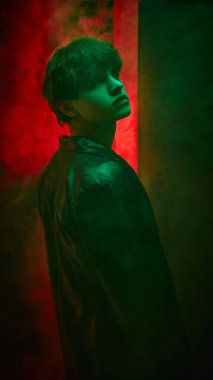 Neon parlak bir portre. Deri ceketli genç adam kırmızı siste kaldırılmış çenesiyle koyu yeşil arka plana doğru dönüyor. Güzellik ve moda kavramı, kendini ifade etme, insani duygular.