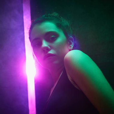 Saçı topuzlu genç bir kadın pembe ve yeşil ışıkların altında dikey parıltılı siyah duvara bakıyor. Etkileyici bir stüdyo resmi. Güzellik ve moda kavramı, kendini ifade etme, insani duygular.