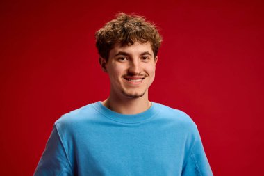 Gülümseyen genç bir adamın portresi. Kıvırcık saçlı, kırmızı arka planda mavi kazak giyen neşeli bir öğrenci. Güzellik, moda, tarz, gençlik, yaşam tarzı, refah, pozitiflik kavramı.