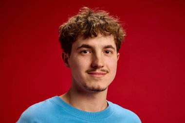 Genç adamın yakın plan portresi, kıvırcık saç stili, hafif bir gülümseme, koyu kırmızı arka planda günlük mavi gömlek giyiyor. Güzellik, moda, tarz, gençlik, güven, ifade.