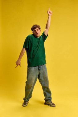 Sıradan yeşil gömlekli ve kot pantolonlu şık bir adam ifade edici yüzüyle dans ediyor ve sarı stüdyo arka planına karşı kollarını kaldırıyor. Gençlik kavramı, hareket, duygu, güzellik, tarz, müzik, eğlence.