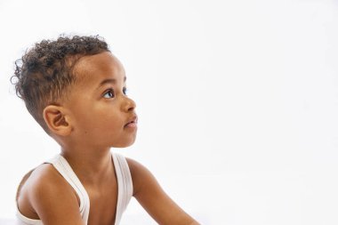 Beyaz kolsuz tişörtlü meraklı çocuğun yakın plan fotoğrafı beyaz stüdyo arka planına karşı negatif arkaplanlı sakin bir ifadeyle yukarı bakıyor. Güzellik kavramı, çocukluk, ebeveynlik, sağlık..