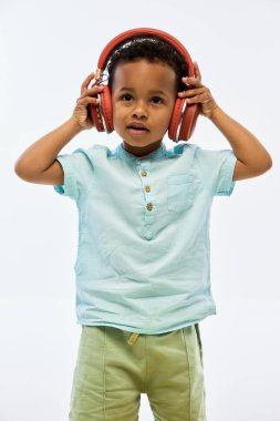 Minyon gömlekli çocuk büyük kırmızı kulaklık takıyor ve beyaz stüdyo arka planına karşı dikkatle dinliyor. Çocukluk kavramı, teknoloji, müzik, dinleme, öğrenme, bakım.
